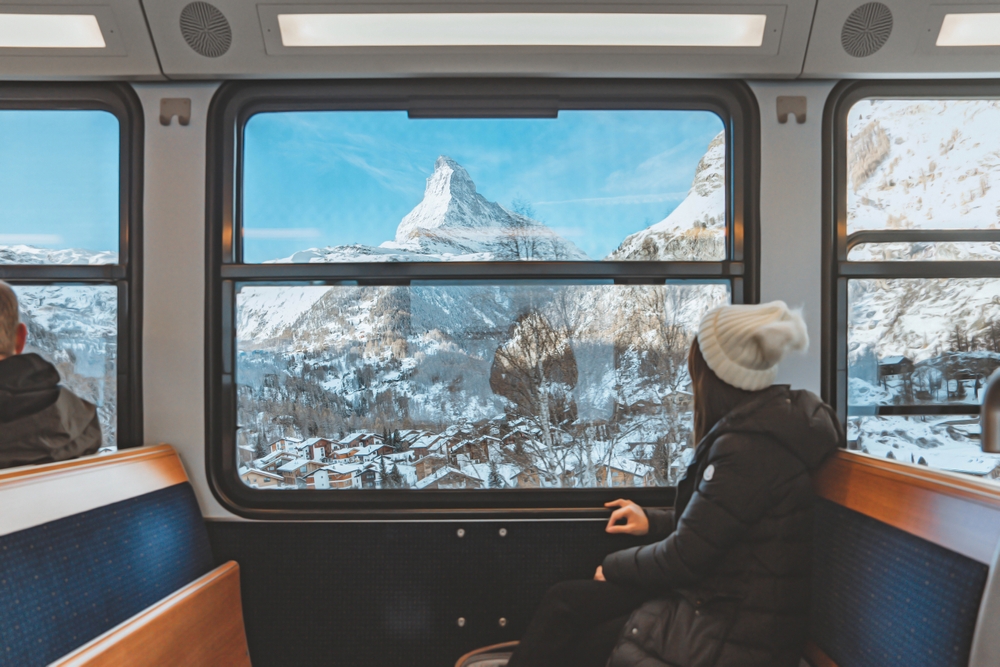 2250519361 Blur foreground - young woman traveling looking out the window enjoying in Swiss Alps with the Matterhorn in winter background while sitting in the train. Tourist travel concept.